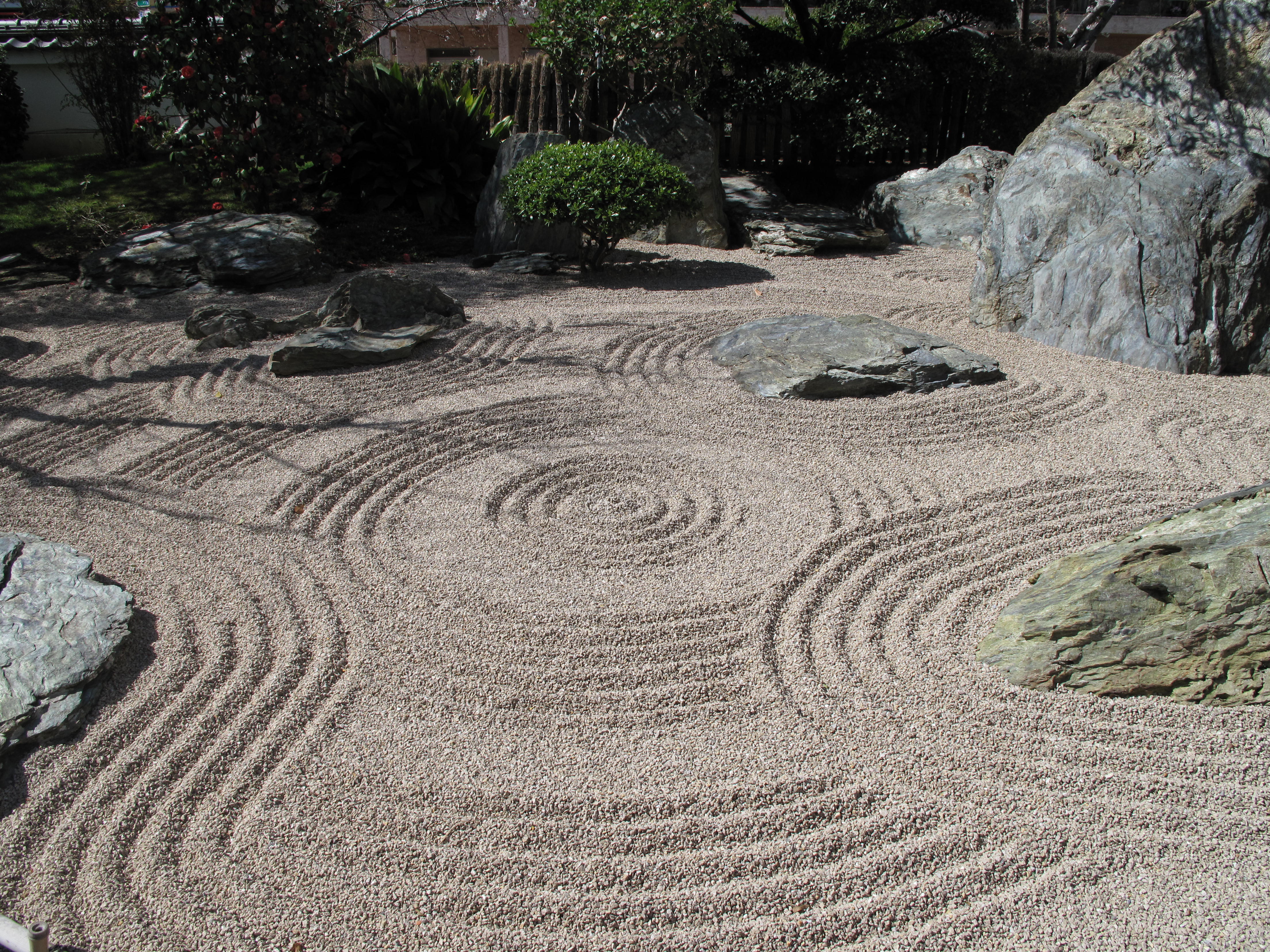 A zen garden of rocks and shrubs surrounding sand that has been spread out in pretty, swirly patterns