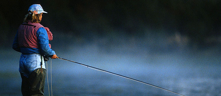 A woman fly fishing in a misty river in dim light