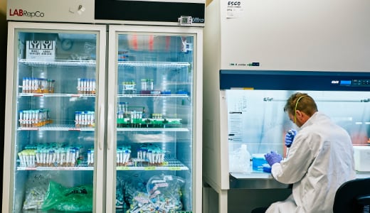 A person in a laboratory wearing a white coat works at a table next to a cooler full of test samples.