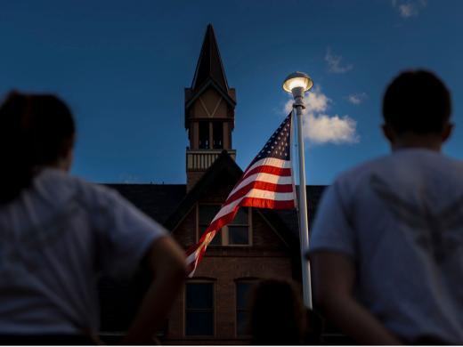 ROTC cadets stand facing an American flag on a pole in front of a college academic building. 