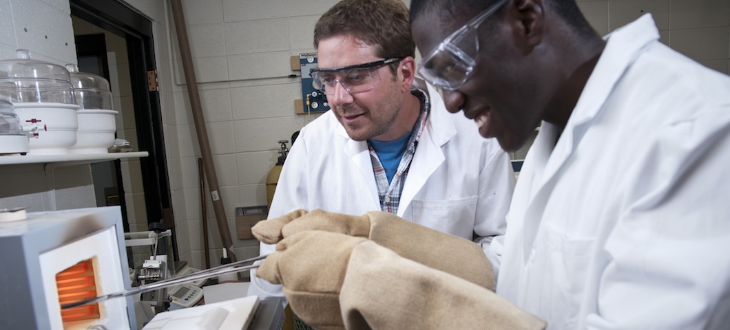 Two male researchers collaborate as one uses tongs to put something in hot oven.