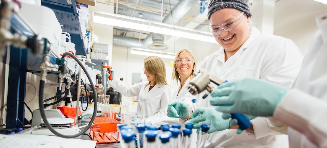 Three women in white lab coats conducting research in lab.