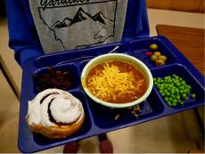 A photo of a student holding a lunch tray with chili, vegetables, and homemade cinnamon rolls