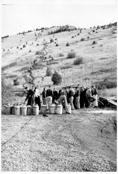 Group of students gathered around milk cans preparing whitewash. Other students are ascending Mount Baldy in background.
