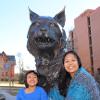 Elva and her daughter Rebecca smile while sitting in front of the bobcat statue with Leon Johnson and Montana Halls in the background