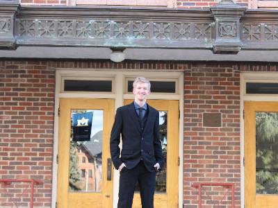 Wade is in a black suit standing outside of a brick building. The door has a reflection of the MSU banner.