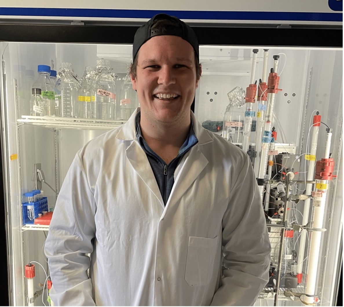 Trevor happily smiling at the camera while standing nonchalantly in front of a bunch of expensive lab equipment